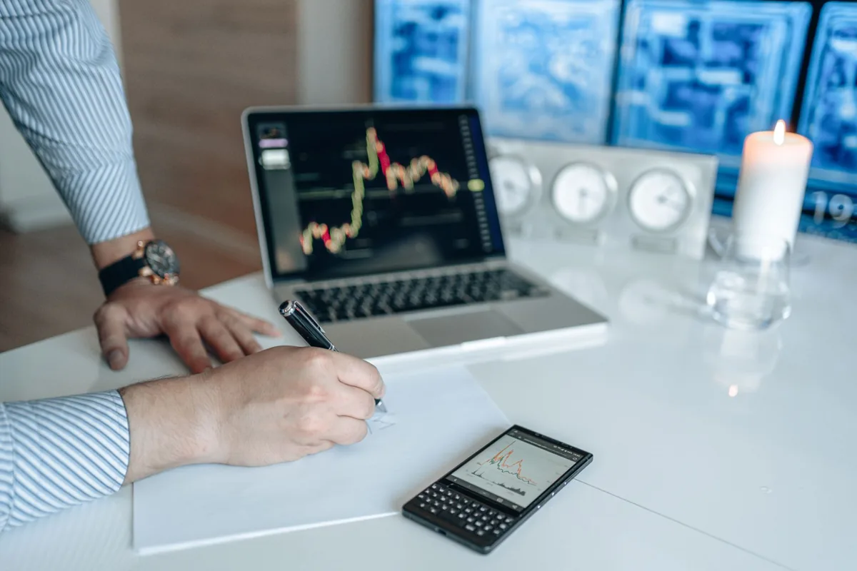 Trader writing pre-trade notes on paper next to a laptop showing a stock chart